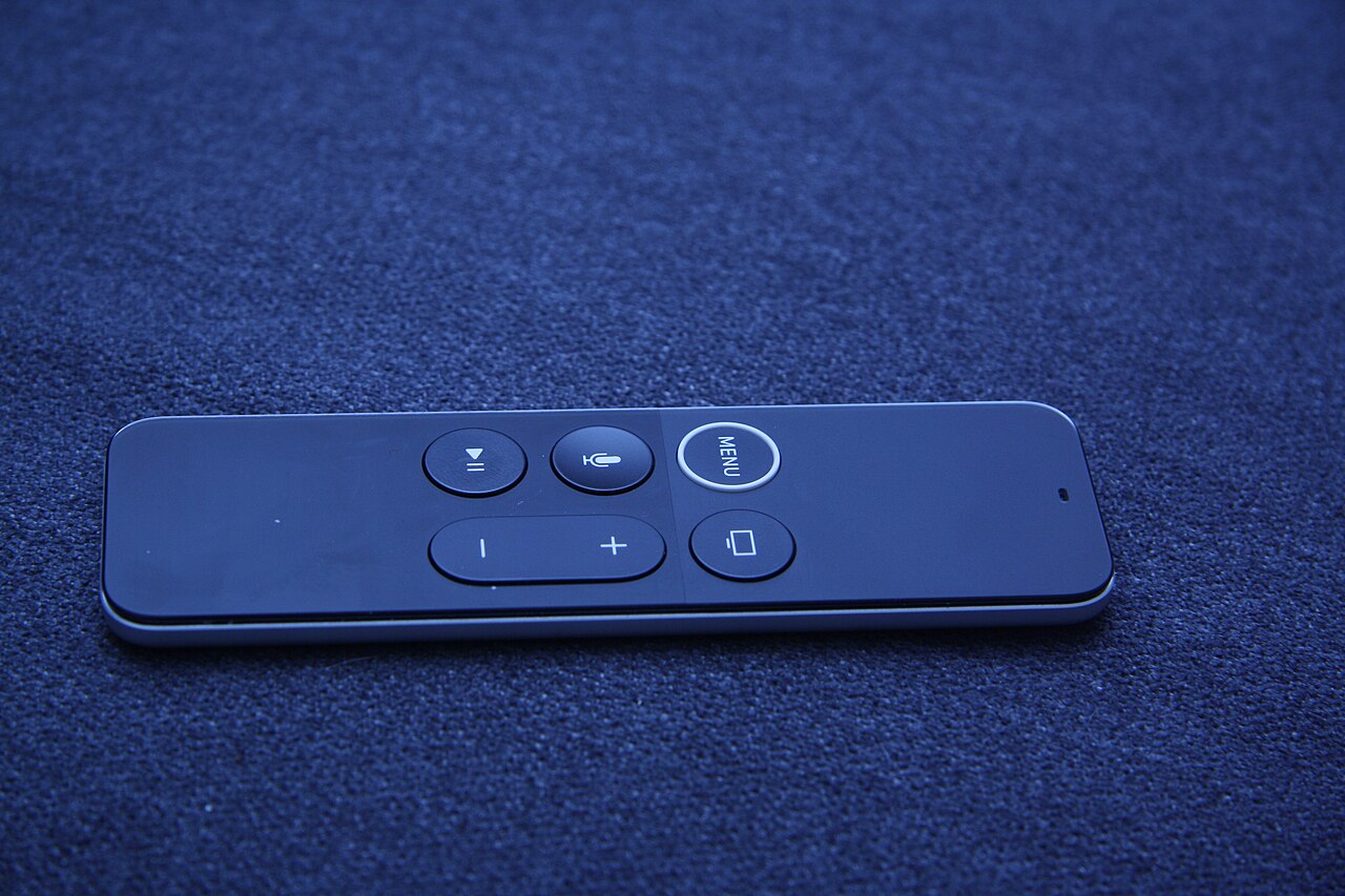 A black first-generation Siri Remote with a white ring around the Menu button, on blue fabric.